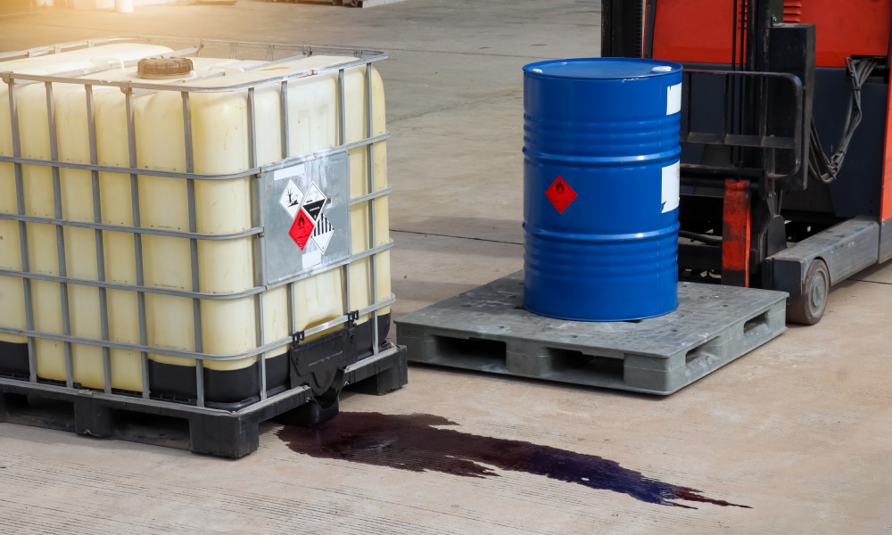 A forklift carries a drum of hazardous materials, while another storage tank spills dark liquid on the concrete floor.