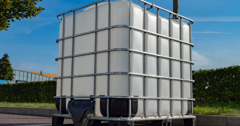 A white plastic tank storing liquid sits in a parking lot. There's a metal frame around the tank and trees in the background.