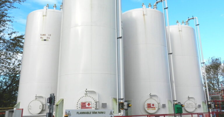 Four large white industrial storage tanks stand tall against a deep blue sky in the background with clouds and trees.