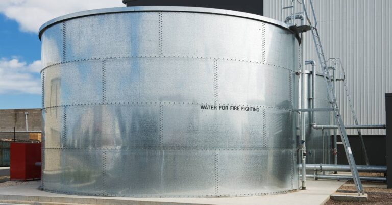 A large steel water storage tank sits by a parking lot with the words "WATER FOR FIRE FIGHTING" on the side.
