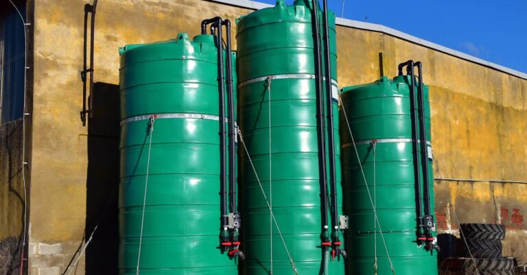 Three green silos of different sizes sit on a platform against a tall wall. They each have pipes connected to them.