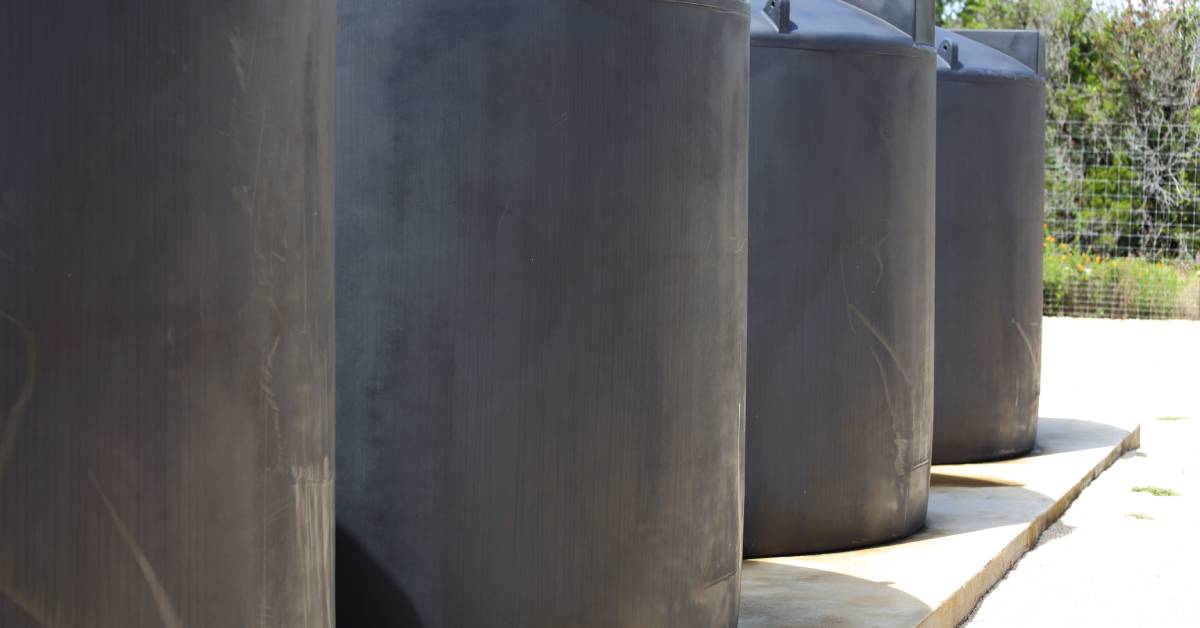 Four concrete water tanks are all lined up next to each other, sitting on a concrete slab surrounded by a fence.