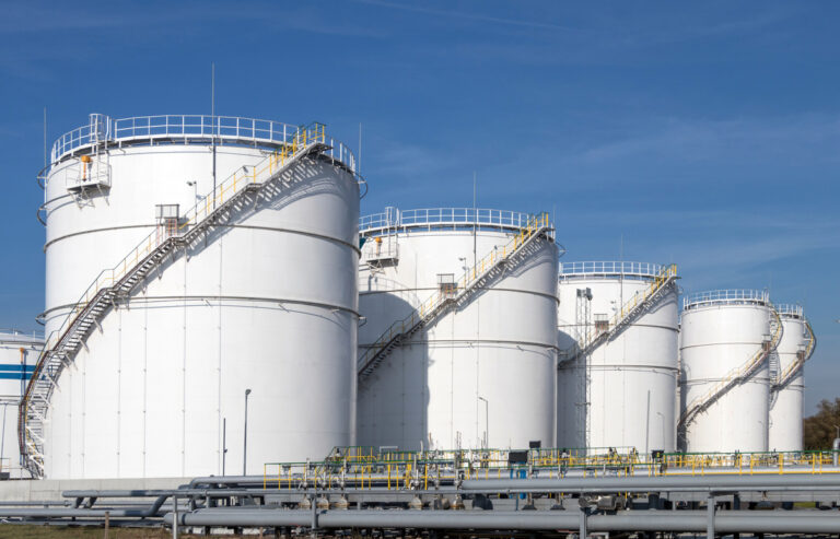 Large storage tanks sit with staircases going around them in a circular pattern. The sky is blue behind them.
