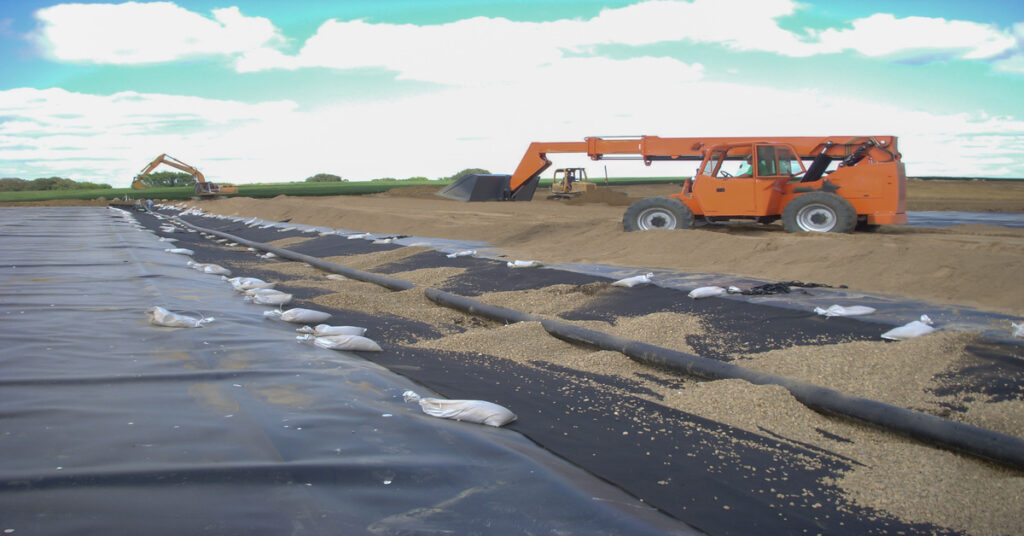 A large machine in the distance moves material around a trench and liner with a pipe down the trench.