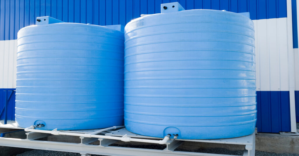 Two large storage tanks sit on top of a structure while the building behind the tanks are striped blue and white.