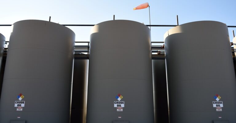 Three gray crude oil storage tanks stand next to each other in the sun, connected by some dark pipes.