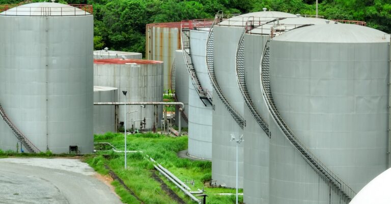Large gray gas storage tanks stand next to each other surrounded by green trees and a grassy landscape.