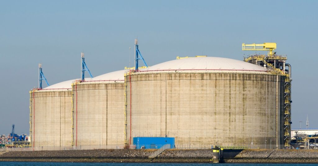 Three large storage tanks stand next to each other near a large body of water. The tanks are off-white.