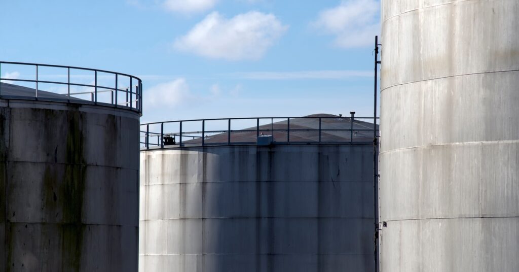Some tall storage tanks standing next to each other. The tanks are made of concrete, and there is a blue sky with some clouds in the background.