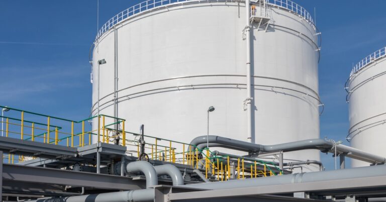 Two large white circular storage tanks next to each other. They're sitting behind a complicated network of pipes and railings,