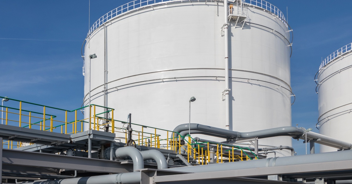 Two large white circular storage tanks next to each other. They're sitting behind a complicated network of pipes and railings,
