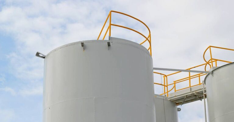 A large white storage tank standing next to other storage tanks, connected by small pipes and yellow railing.