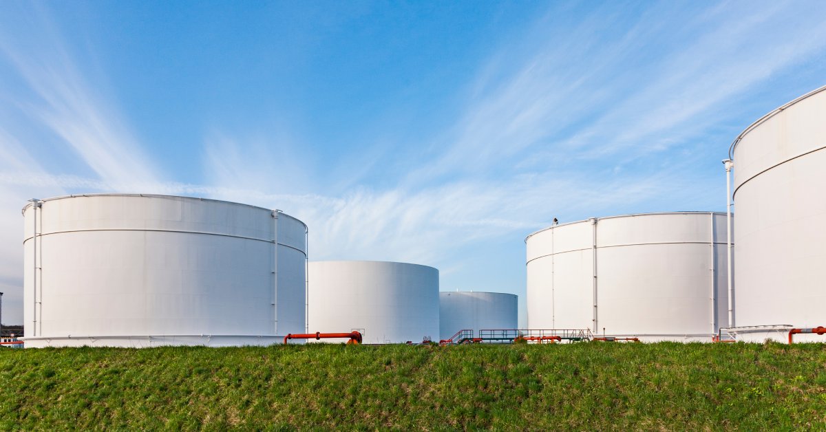 In a large field, multiple large, cylindrical storage tanks stand next to each other with a blue sky and clouds behind them.