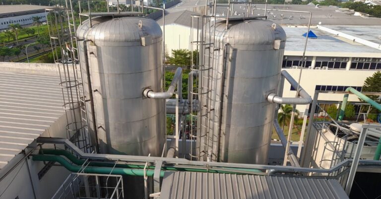 Two large storage tanks stand next to each other with numerous pipes and ladders leading up to the top.