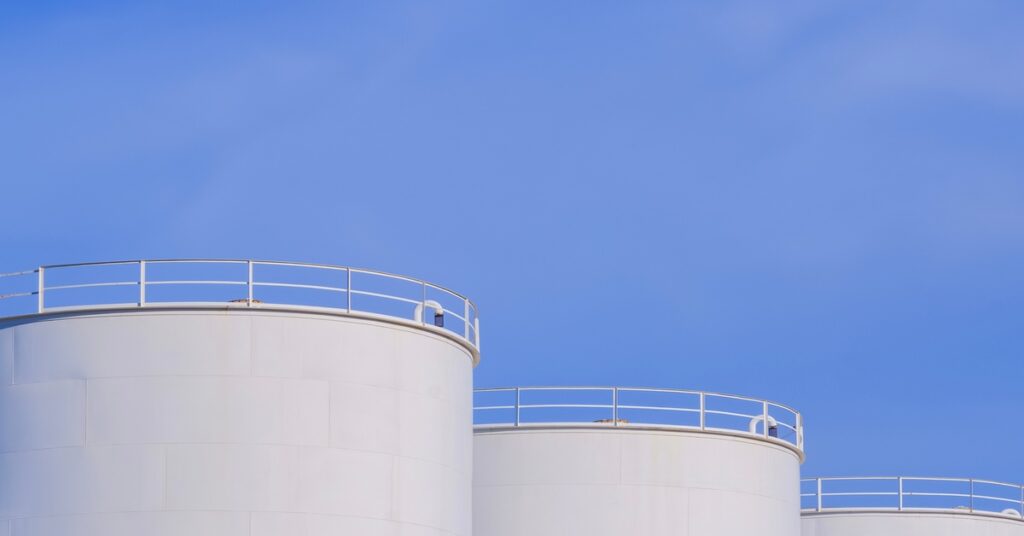 Three large white storage tanks stand next to each other on a concrete platform with a walkway connecting them.