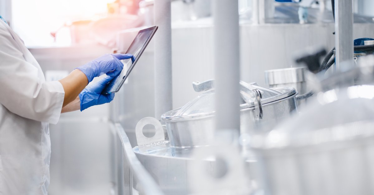 A focus on a food processing worker's hands as they hold a tablet and inspect the metal tanks in a facility.