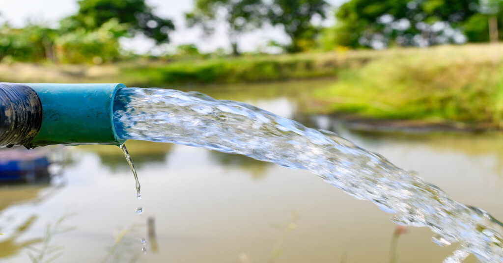 A close-up of a drain pipe with clear water gushing out into a reservoir with green grass in the background.