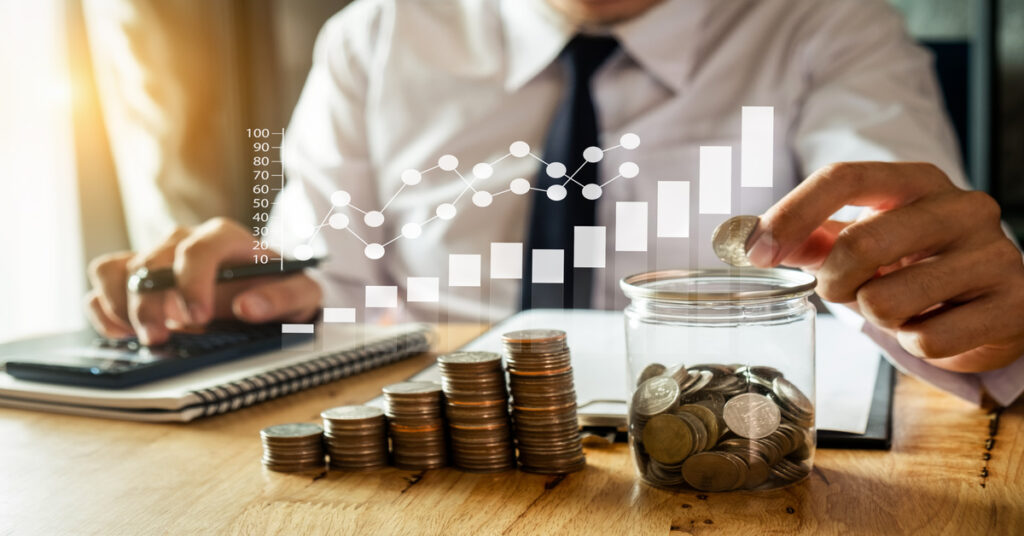 A businessman in a suit and tie sitting at a desk, depositing coins into a jar with an abstract graph floating above.