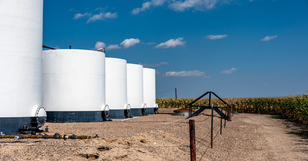 Four large, white containment storage tanks with secondary containment and fencing in a rural corn field.
