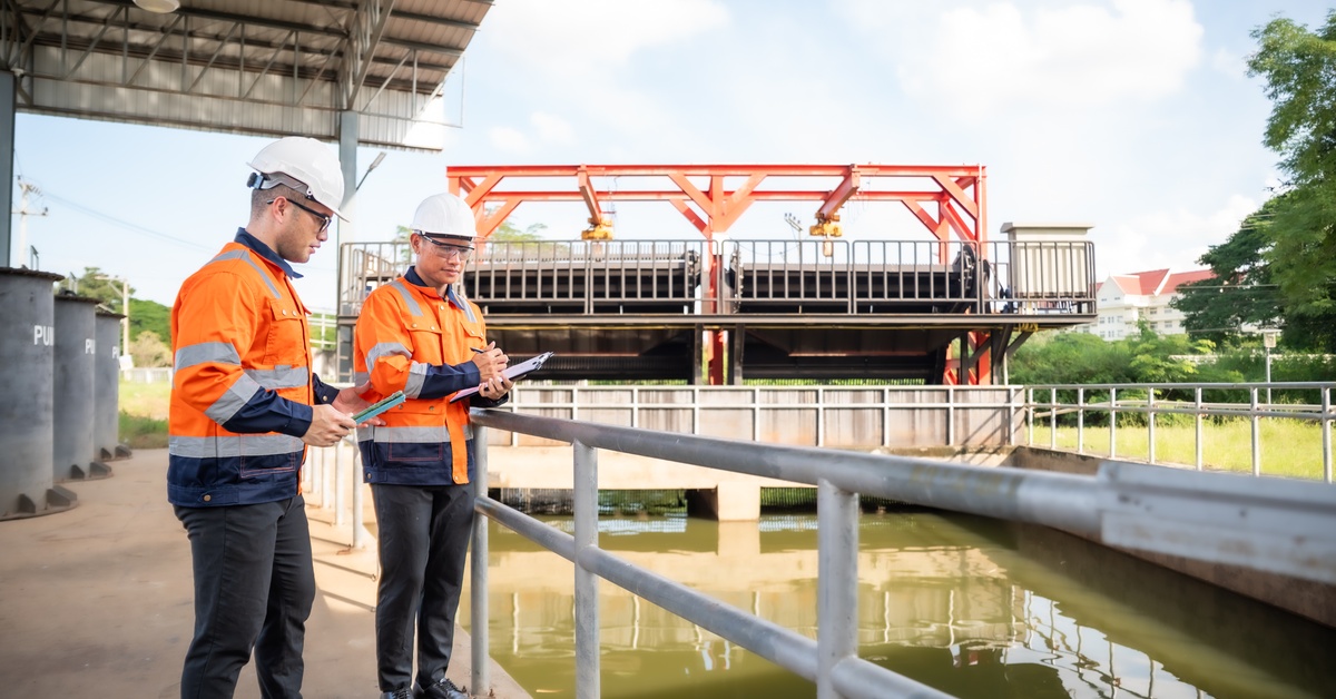 Two men at a water treatment plant wearing safety gear stand with clipboards at a railing near a water tank.