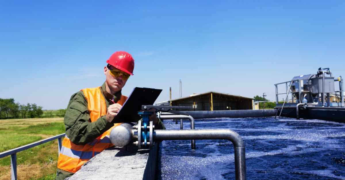 An industrial worker wearing an orange vest and red hard hat holds a clipboard next to a water treatment tank.