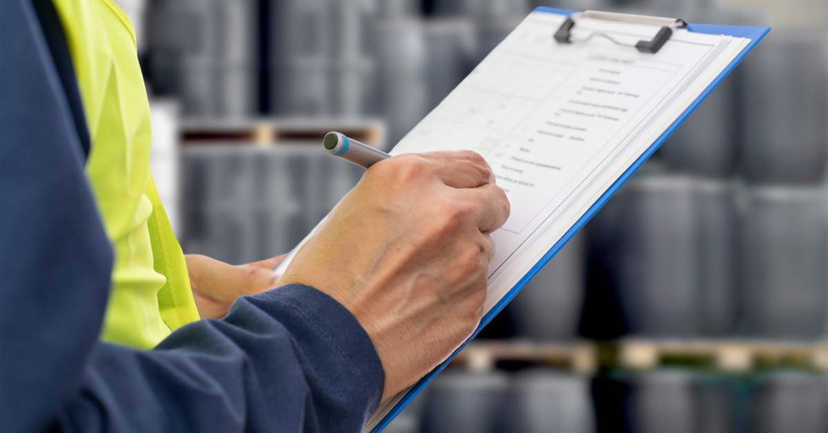 A man wearing a safety vest holds a clipboard and takes notes in a facility or warehouse with containers in the background.