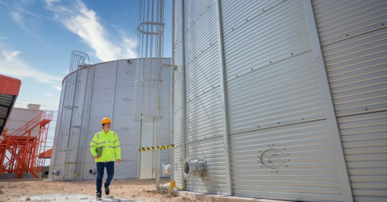 A person in a yellow safety jacket and hard hat walking between tall metal industrial tanks under a blue sky.