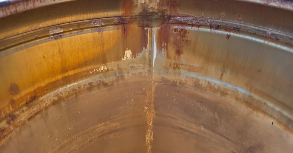 A stained metal storage tank interior with brown residue, mineral buildup, and streaked surfaces along the sides.