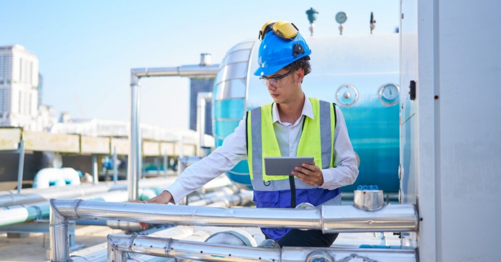 A worker in a blue hard hat using a tablet beside shiny metal pipes and a blue tank at an industrial site.
