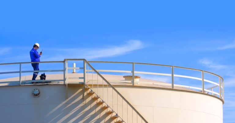 A worker in a hard hat standing on a fuel tank platform while holding a walkie-talkie under a blue sky.