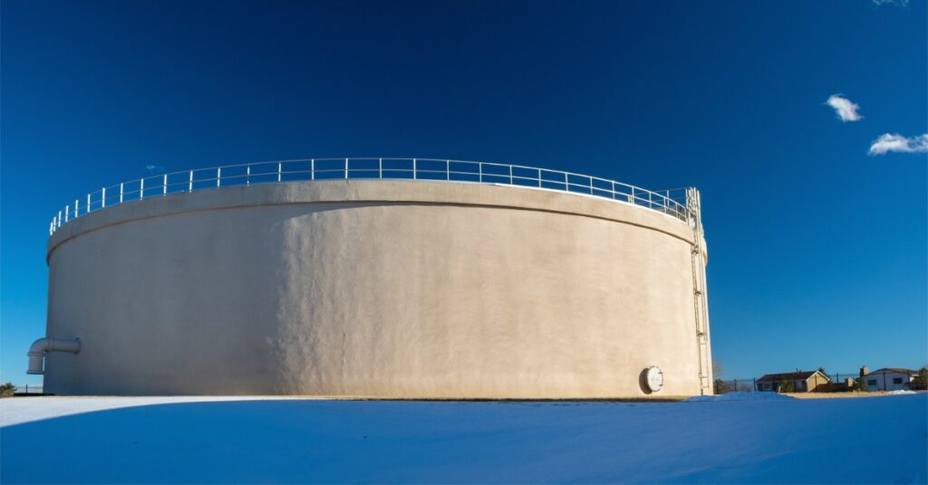A large concrete water tank standing on snowy ground beneath a deep blue sky with small buildings in the distance.