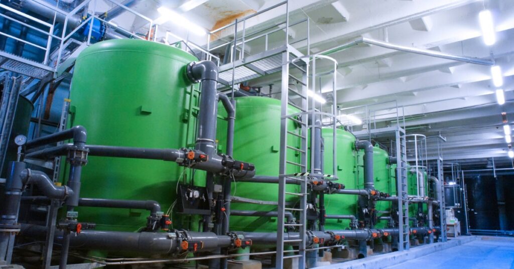 Green cylindrical tanks standing inside an industrial room with gray pipes, ladders, walkways, and bright lights.