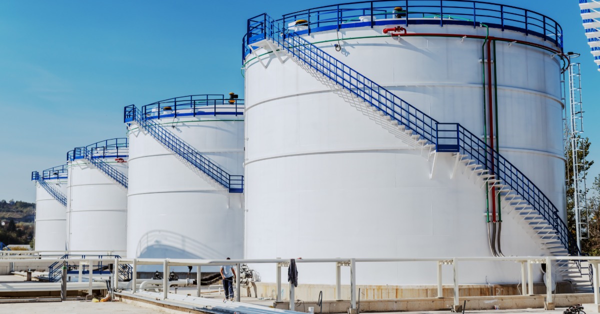 A row of large white cylindrical storage tanks with exterior stairways and railings beneath a clear blue sky.
