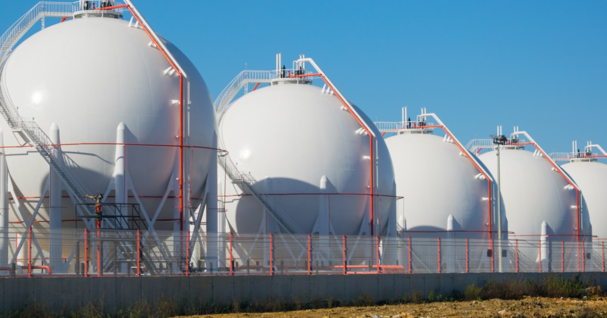 Five large white spherical storage tanks with exterior stairways and red piping standing behind a metal fence.